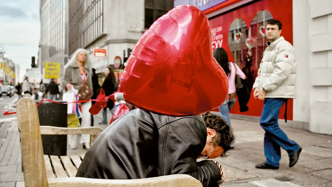 A man sitting on a bench in a shopping street with a large red helium balloon.