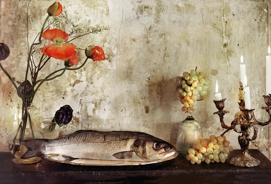 Still-life photograph of a whole fish on a silver platter, surrounded by red poppies, green grapes, glass vessels and lit candles against a textured wall.