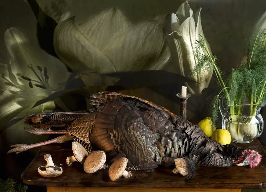 Still life with a wild turkey, mushrooms, quinces, fennel and a candle on a wooden table, set against a dramatic backdrop of oversized white lilies.