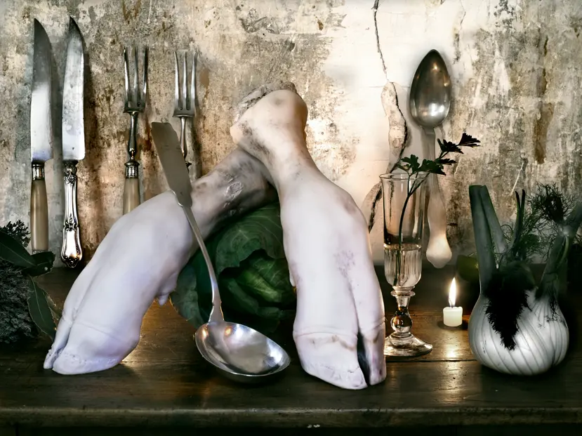 Still life with two pale animal legs arranged over a green cabbage, surrounded by vintage cutlery, fennel, parsley, a glass and a lit candle against a weathered wall.