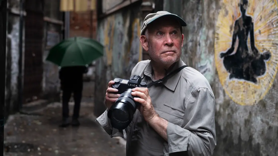 Photographer Steve McCurry holding a Leica camera in a narrow rain-soaked alley in India, with a silhouetted mural and a passer-by carrying an umbrella in the background.