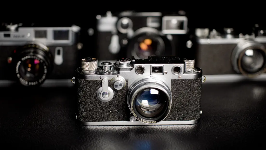 Four Leica Cameras standing on a dark table.