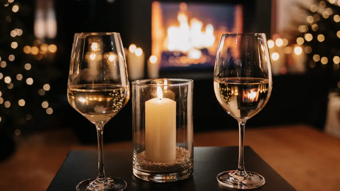 Two filled wine glasses with a candle on the table. A fireplace in the background.