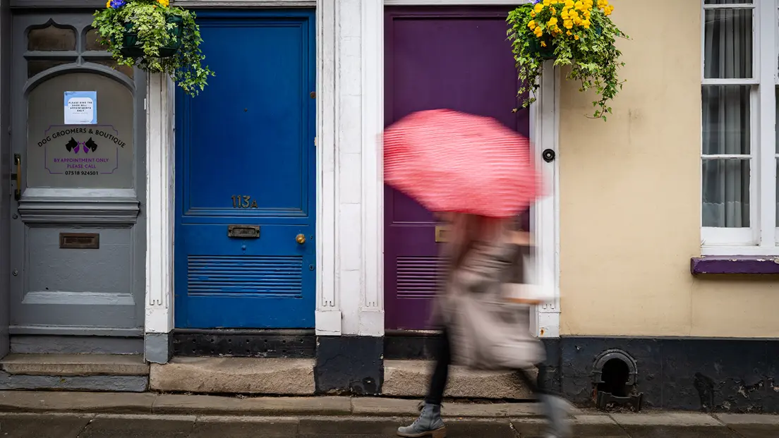 A person is walking past three colorful front doors, holding an umbrella.