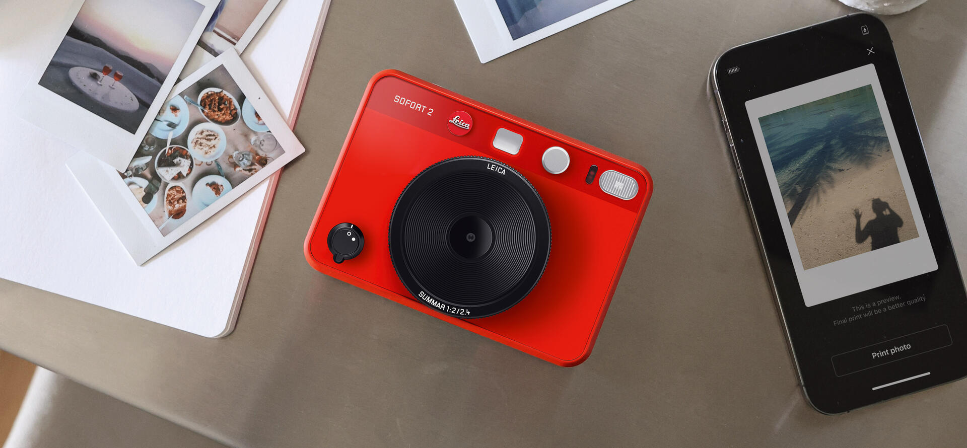 Leica Sofort 2 red in the middle of prints and a mobile phone.