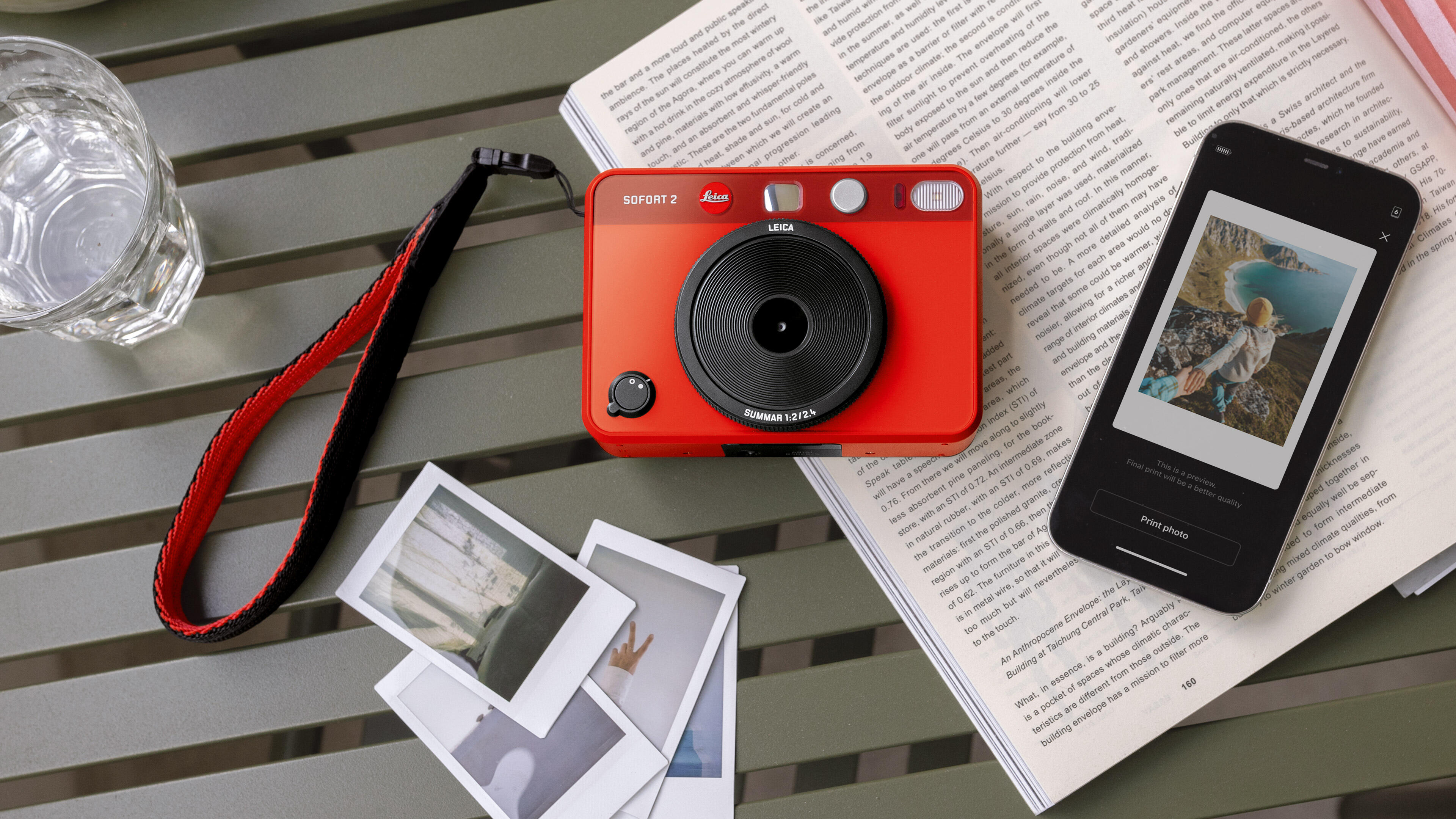 Leica Sofort 2 red, a mobile phone and prints on a table.