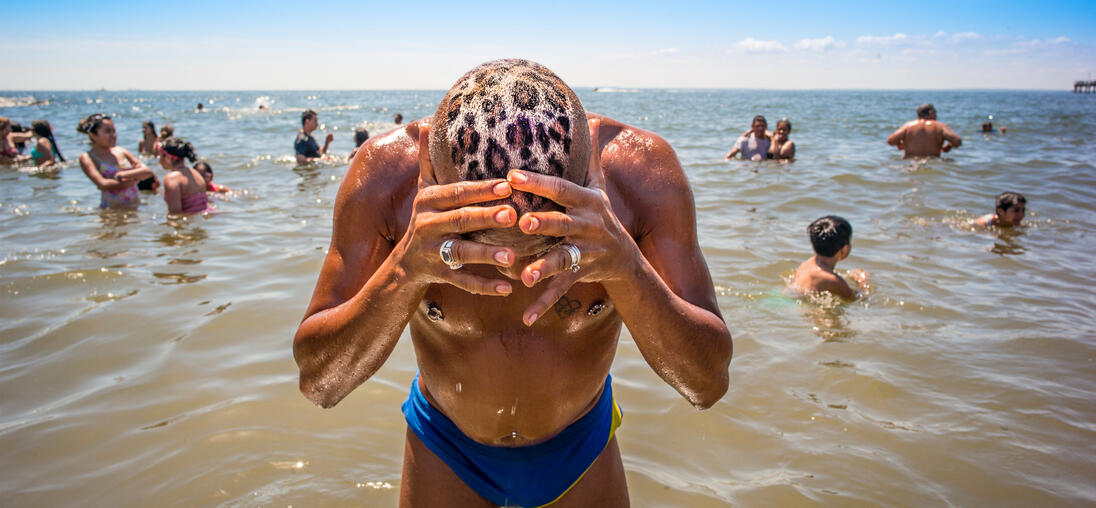 A man with a leopard print on his head is standing in the water. Several people are bathing in the sea around him.