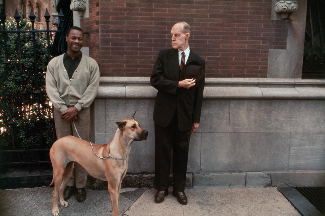 Two men, one with a dog on a lead, stand in front of a brick wall.