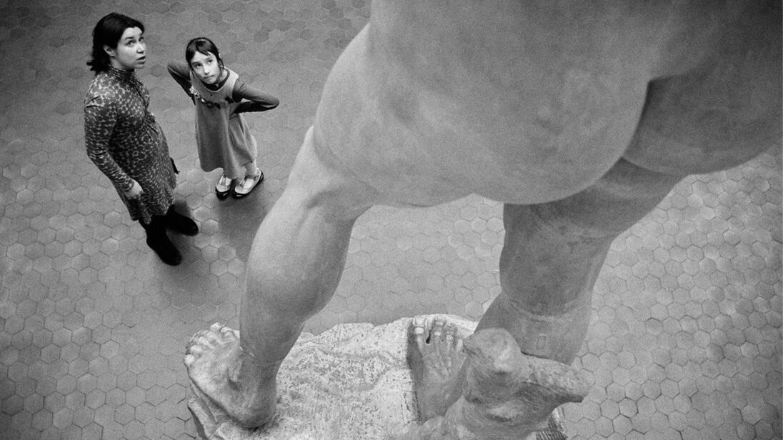 Two people stand in front of a large statue and look upwards.