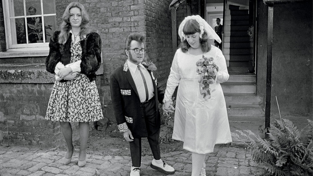 A wedding couple and a woman in a dress stand in front of a building.