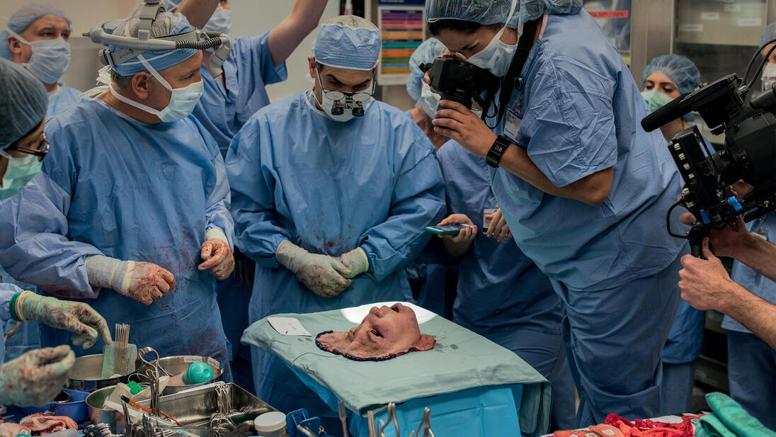 A group of people in surgical gowns look at a mask representing a face, and some take pictures of it.