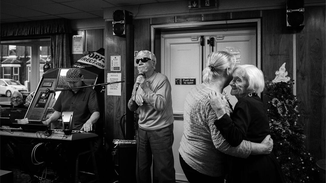 A man sings into a microphone while two older women dance and a man plays the keyboard in the background.