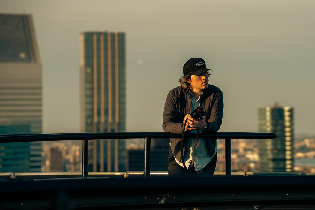 A man holding a camera leans against a railing with skyscrapers in the background.