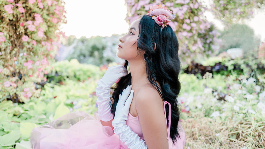 Woman in a pink dress and white gloves is sitting in a rose garden looking to the side.