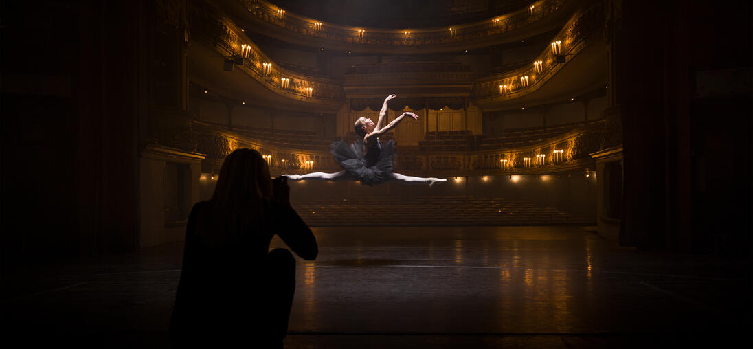 A person in the shade takes a picture of a ballerina jumping in an illuminated theatre.