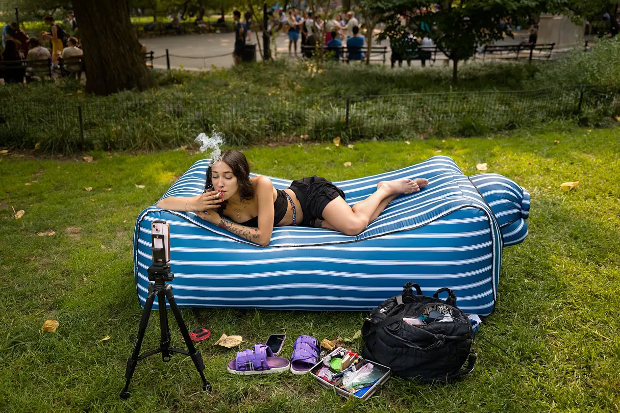 Woman lies smoking in the garden on an air mattress and shoots videos with a mobile phone on a tripod.