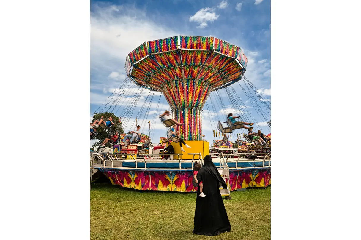 Woman with a child in her arms stands in front of a chain carousel.