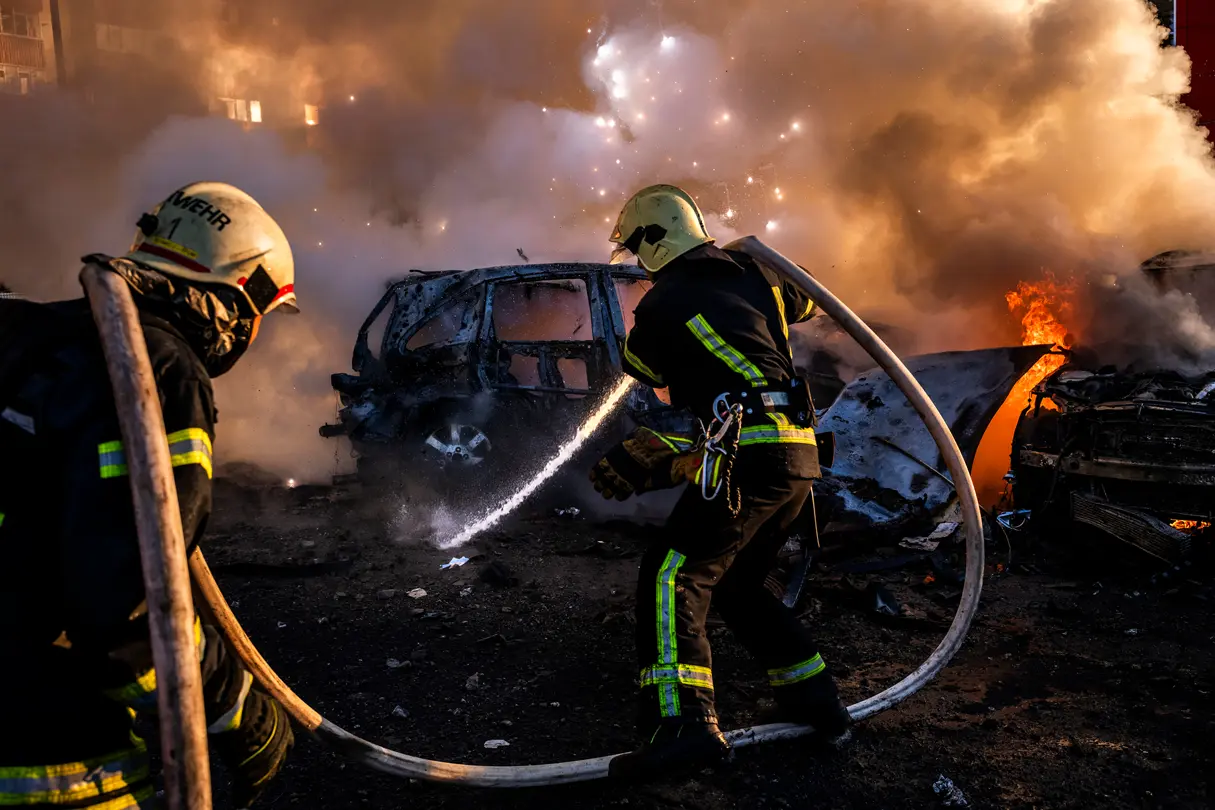 2 firemen extinguishing cars, burning houses in the background