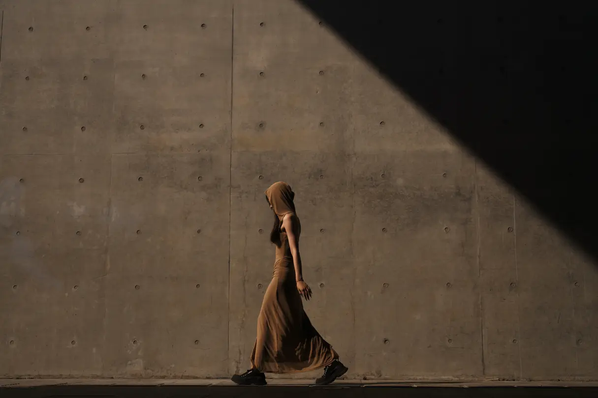 Woman with brown dress, hood and black sneakers walks along a grey concrete wall