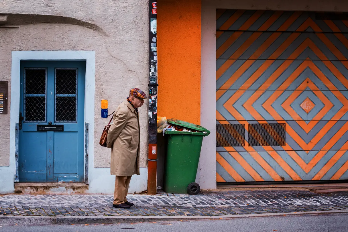Old man standing between a blue door and a full, overflowing green bin