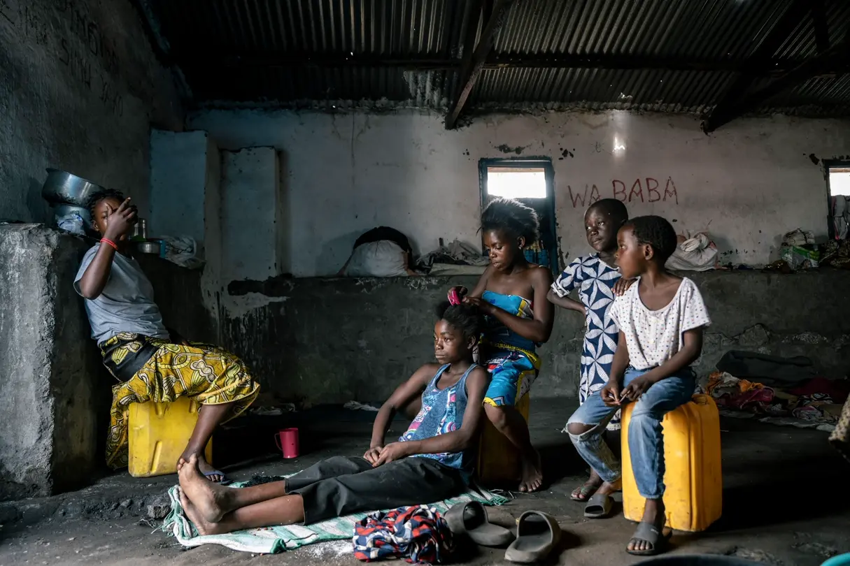 Five children sitting in a run-down hut. One girl braids the other's hair.