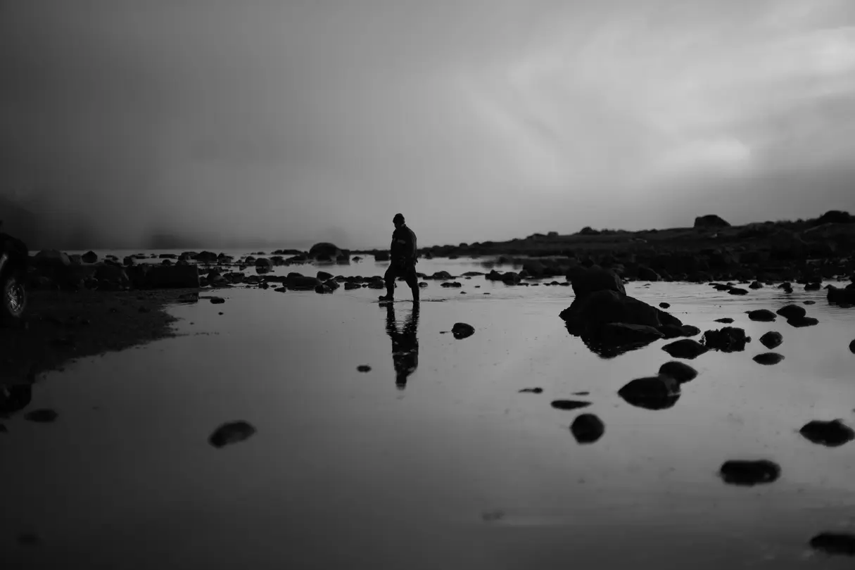 Old man walks through shallow river wearing rubber boots