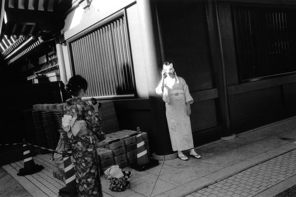 Two Japanese women, one with a camera in her hand, the other holding a mask in front of her face.