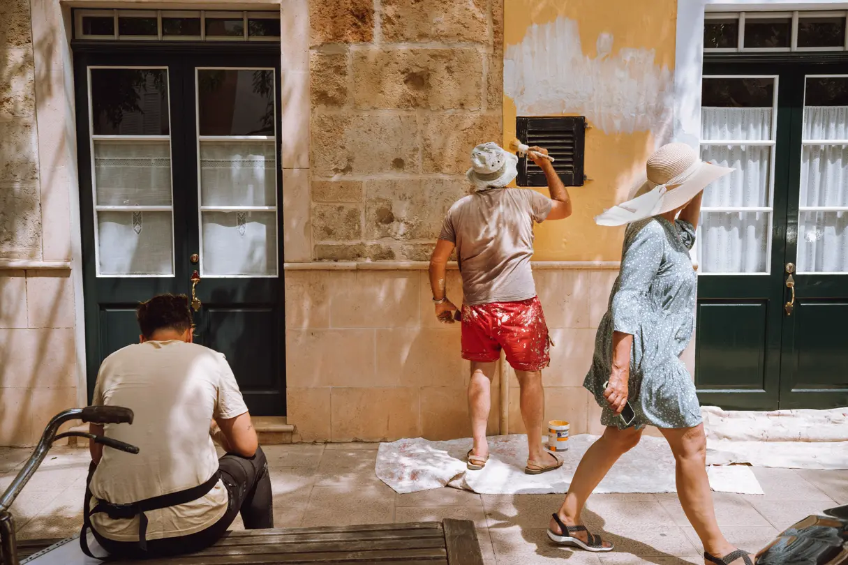 A man sits on a bench while another man paints the wall of a house. A woman walks past, talking on the phone.
