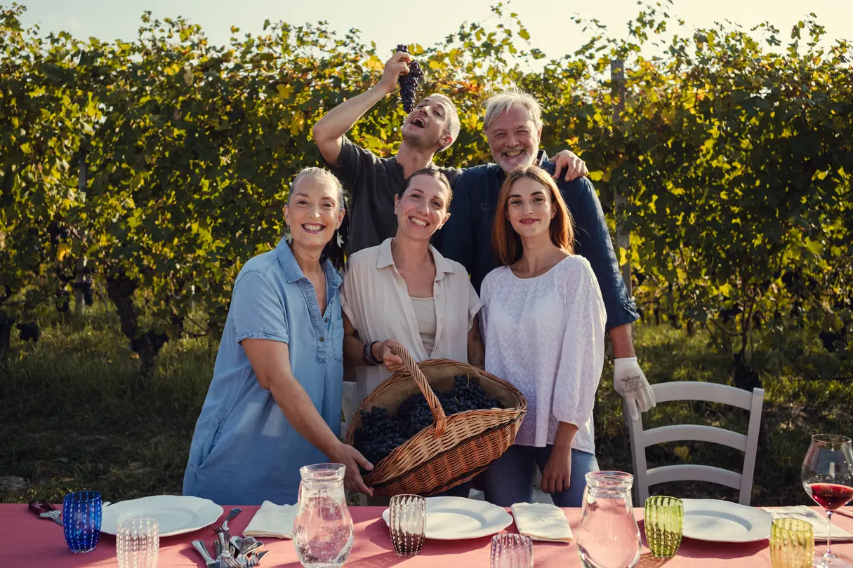 A family stands in front of a laid table in the vineyards, and a woman holds a basket of grapes in her hand.