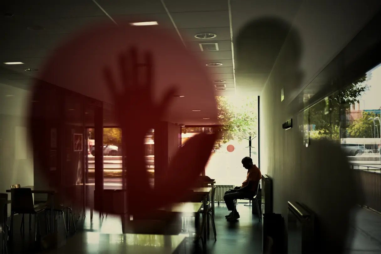 Joaquín Pastor - Chico sentado en una silla en un local y reflejo de otro chico con la mano levantada en una pegativa roja redonda