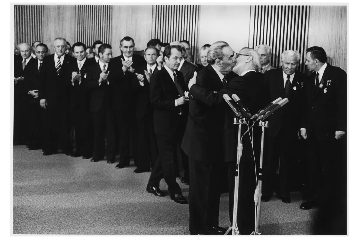 two elderly men in suits kissing in front of a row of other men in suits