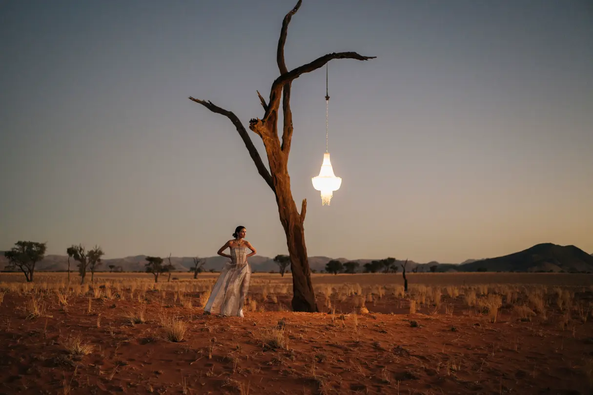 Woman in a dress standing under a tree with a chandelier in the desert at sunset