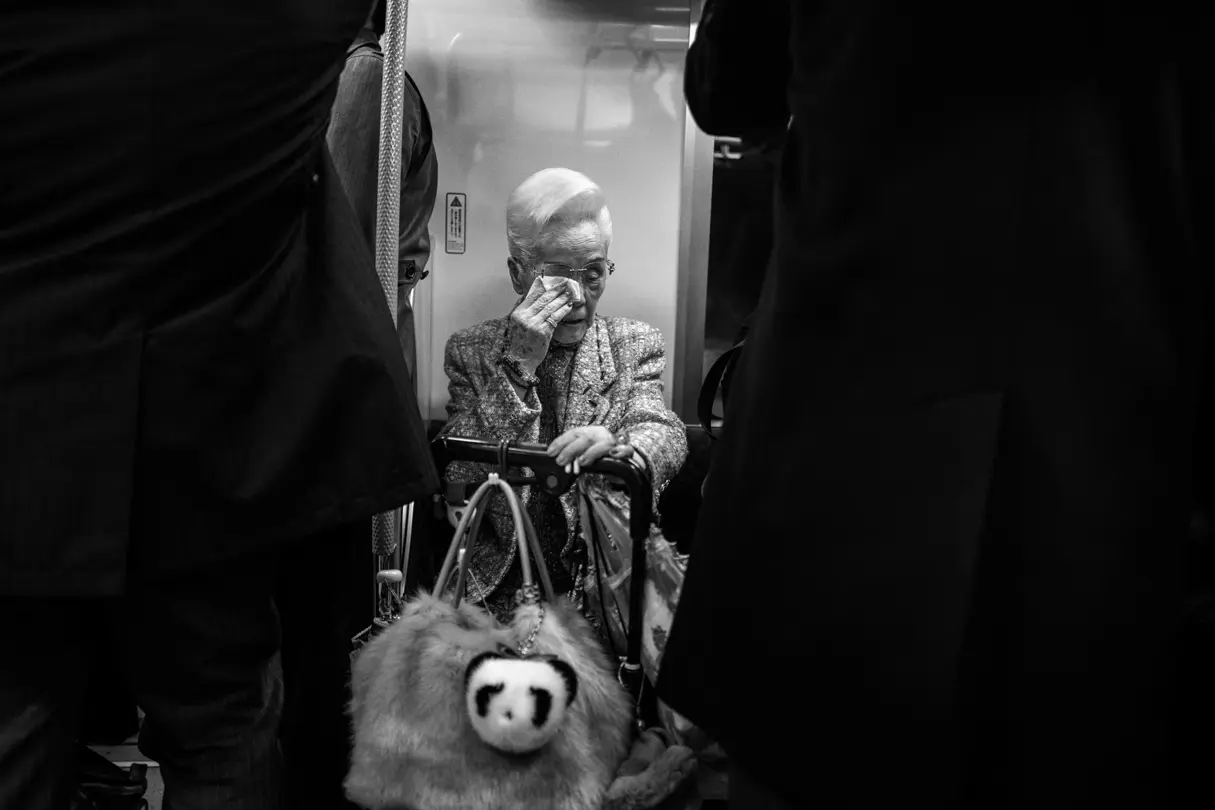 A stylish elderly woman sits in the underground and wipes her eye with a tissue.
