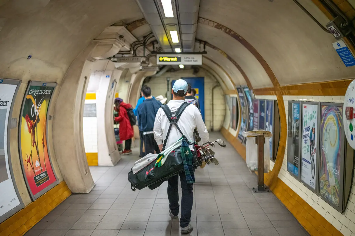 Man in an underground station carrying a large bag with golf clubs slung over his shoulder.