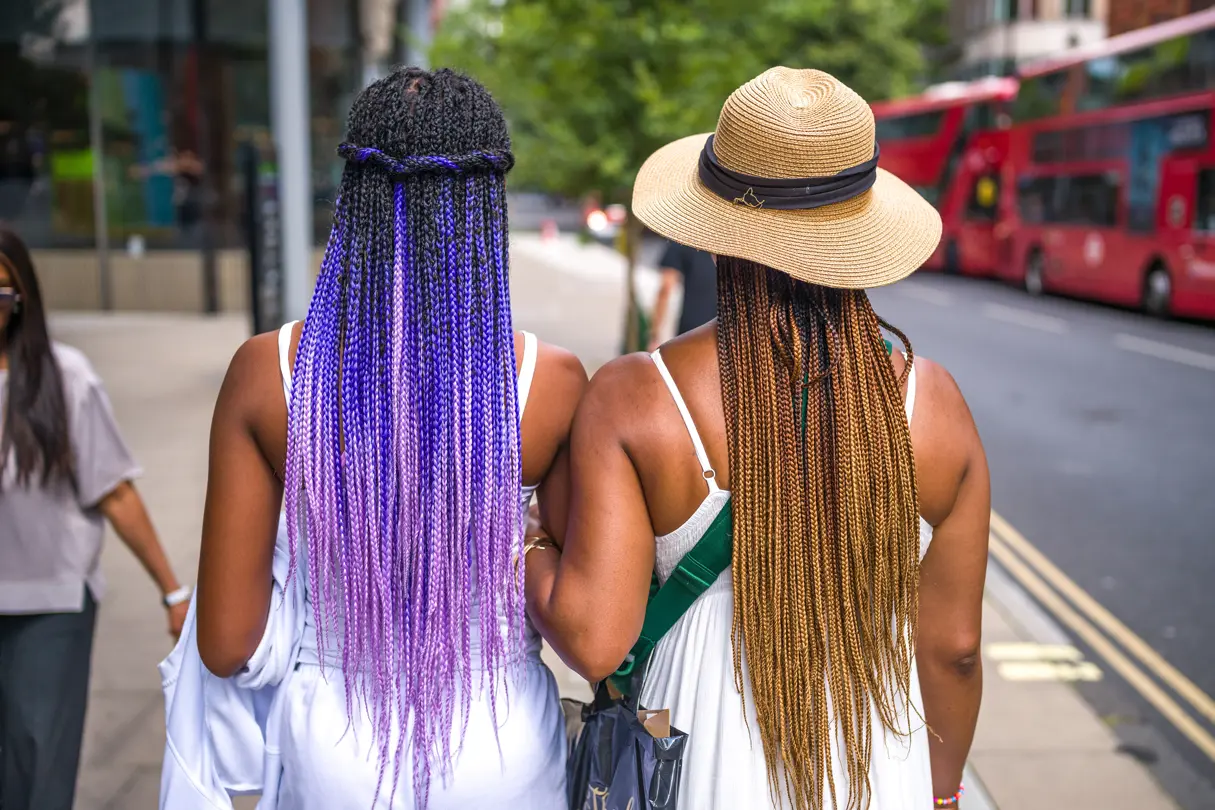 Two women with dreadlocks, one with purple hair and one with brown hair and a hat, photographed from behind.