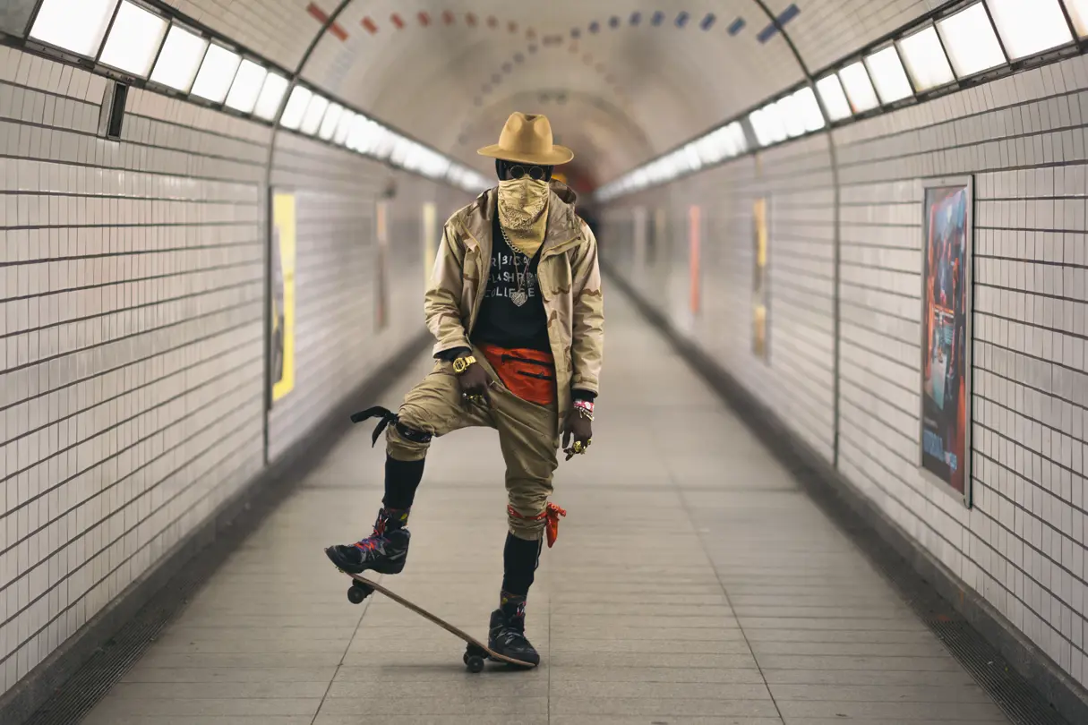Coloured man on a skateboard in an underground station wearing a hat and scarf pulled up over his nose