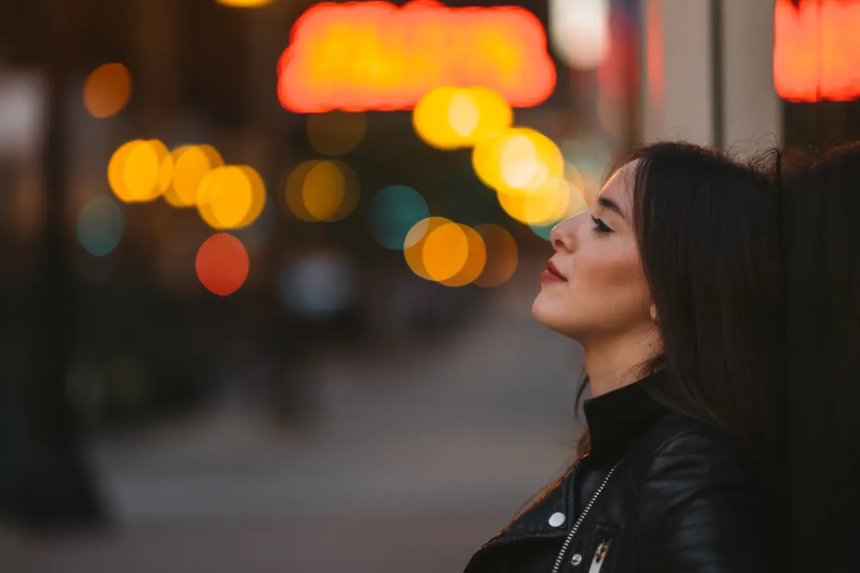 Side portrait of a young woman with dark hair at dusk, with blurred lights in the background.