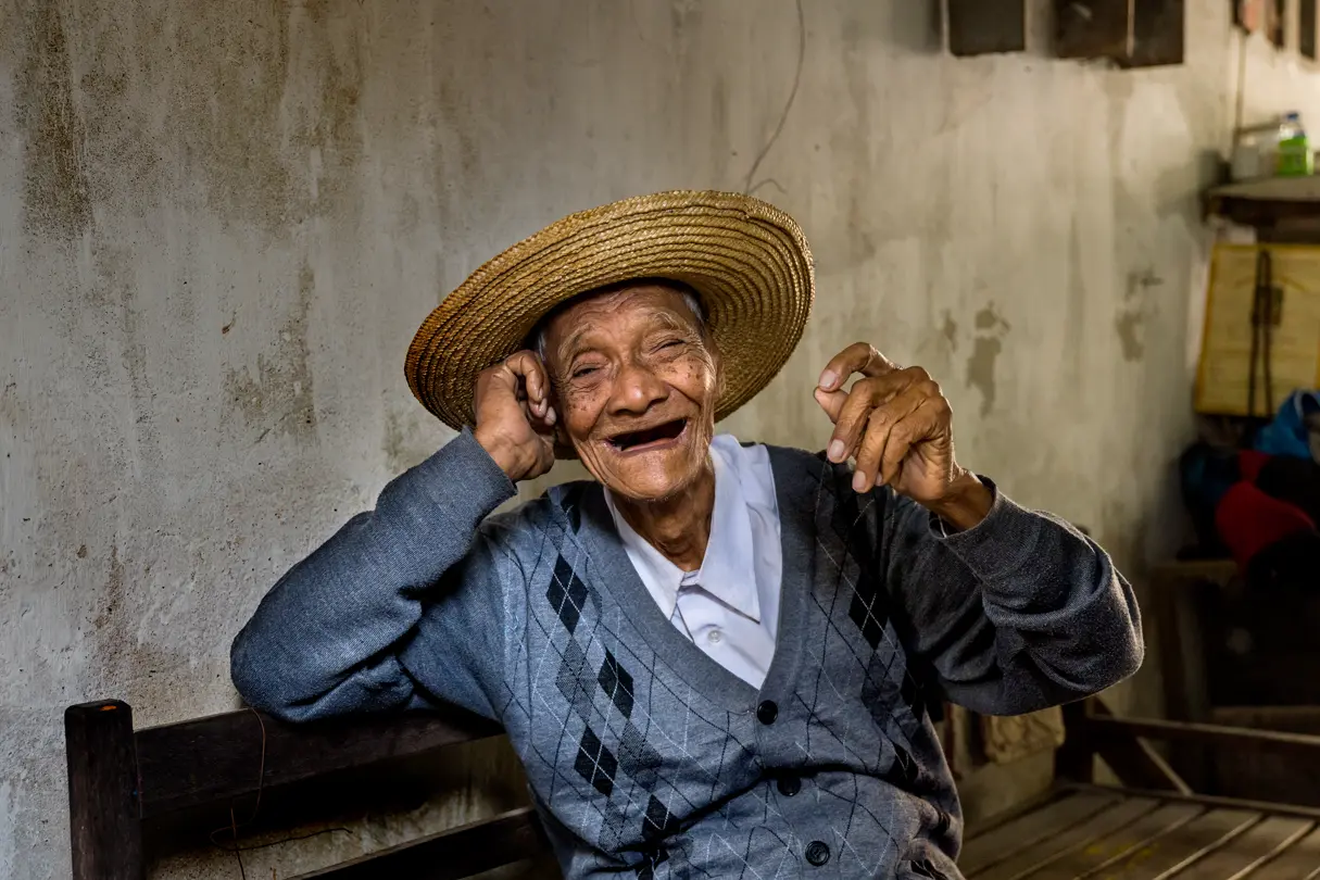 An old man in a large hat laughs at the camera