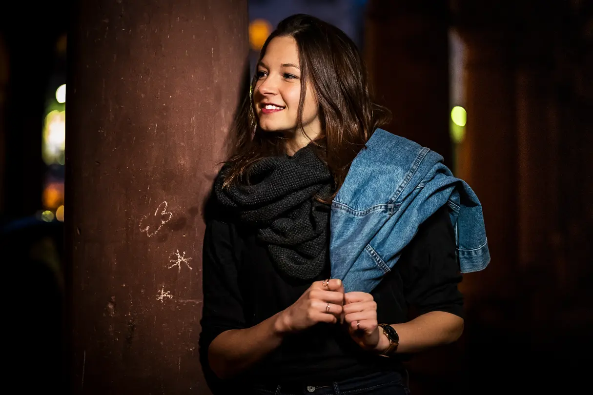 A portrait of a young brunette woman with a denim jacket draped over her shoulder, leaning against a post