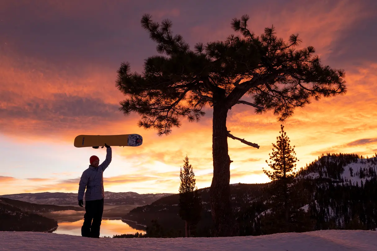 A snowboarder holds his snowboard aloft on a mountain at sunset