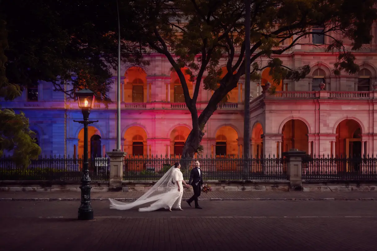 A bride and groom in front of an old building, illuminated in blue and red in the darkness