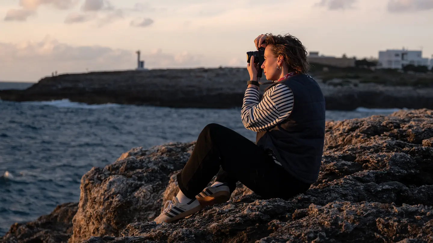 Julia Nimke is sitting on a rock by the sea with her Leica camera, taking photos