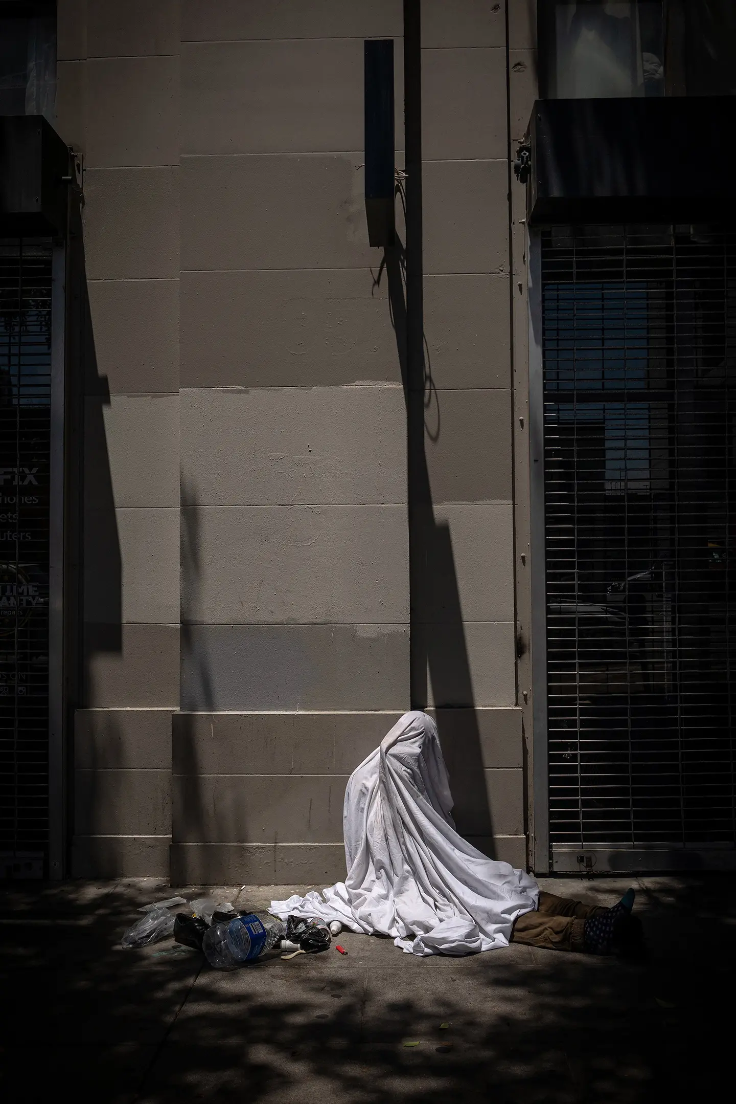 Person resting beneath a white sheet on a city sidewalk, capturing themes of urban life, vulnerability, and social reality.