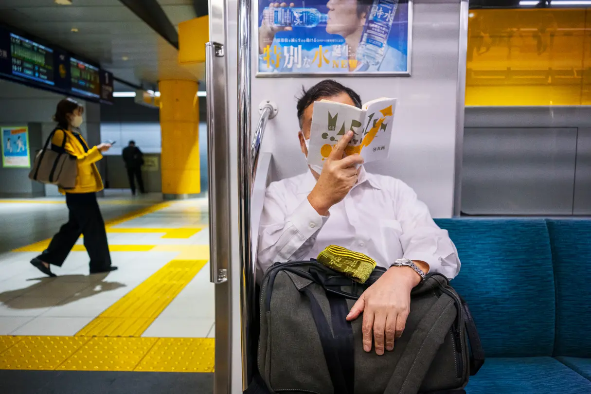 Commuter reading a book on a Tokyo subway train while passengers move through a brightly lit station platform. Candid urban photography capturing everyday routines, public transport, and contemporary city life in Japan.