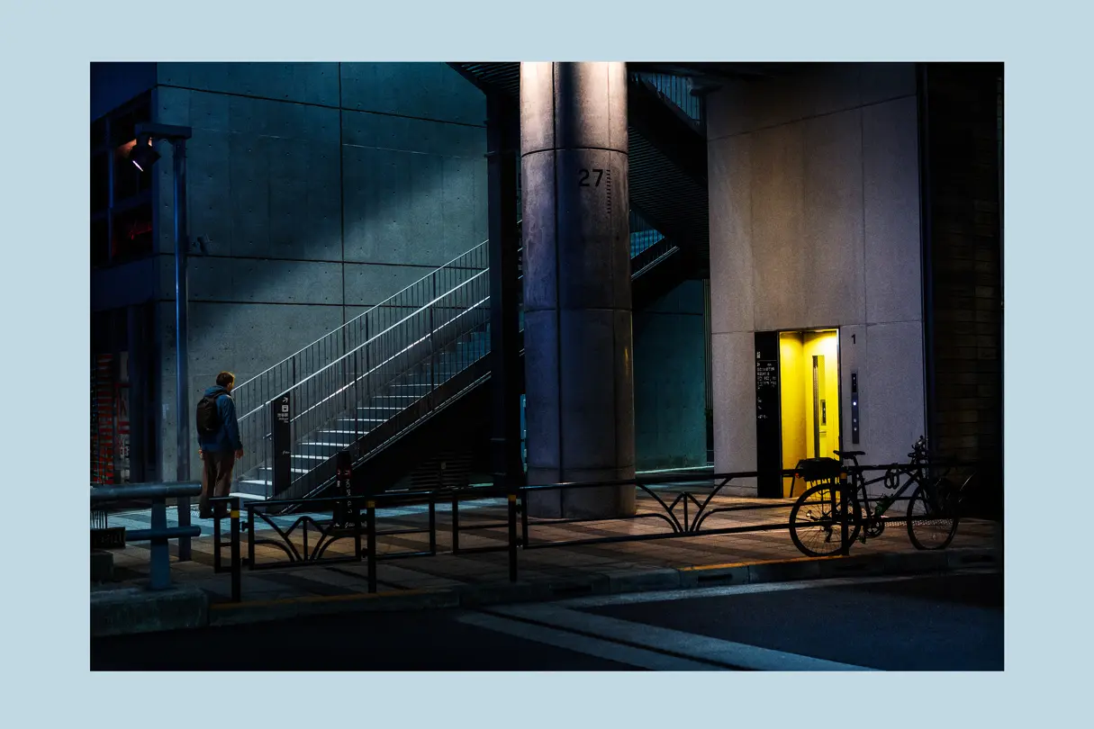 A solitary figure walks through a modern urban space illuminated by cool blue tones and a glowing elevator. Contemporary Tokyo street photography with strong geometric composition.