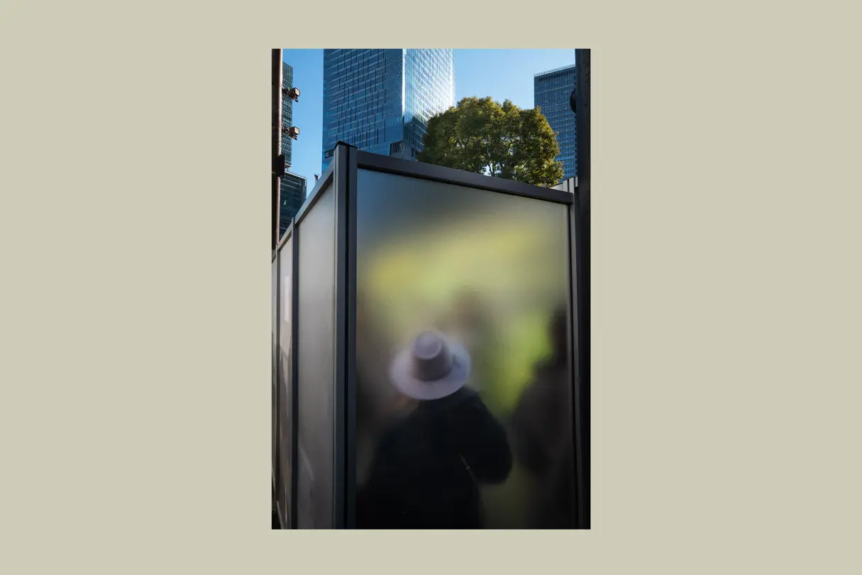 Blurred silhouette behind a translucent city structure framed by modern architecture. Abstract street photography exploring perception, anonymity, and urban life.