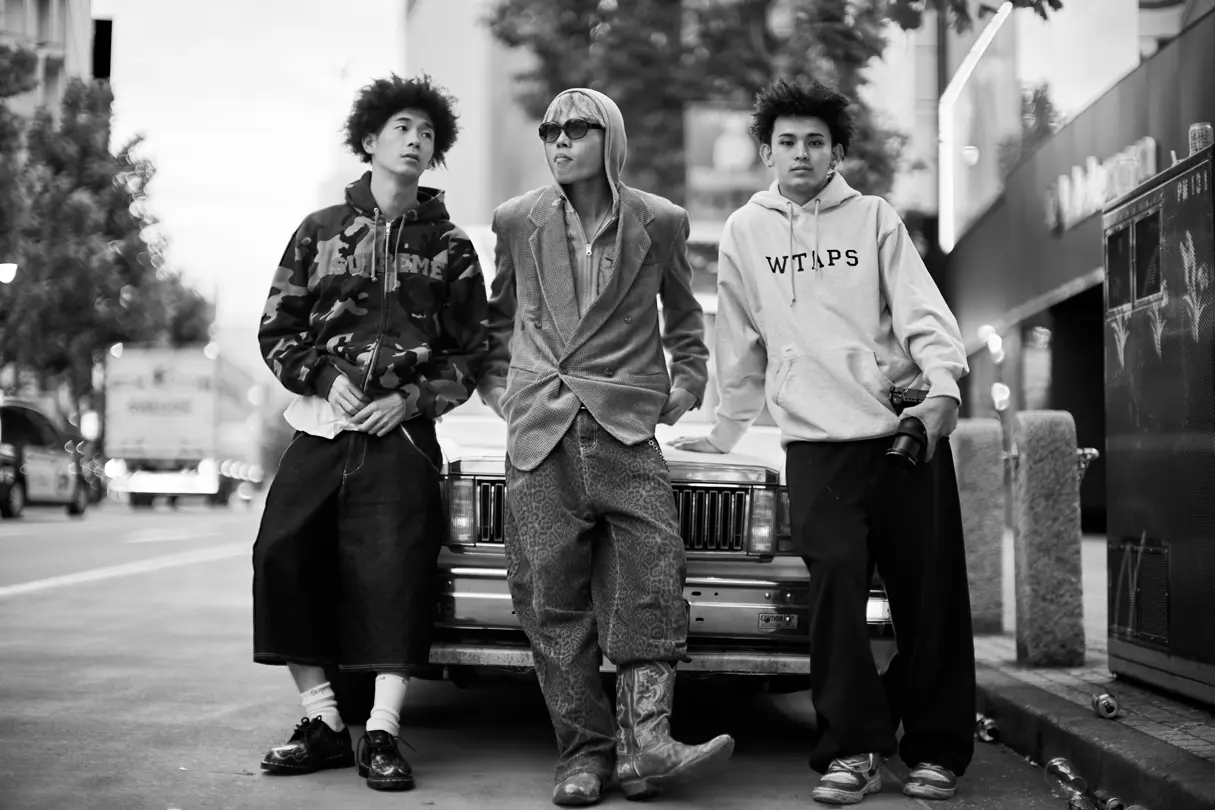 Three young creatives pose beside a vintage car in Tokyo, showcasing contemporary street fashion in black and white.