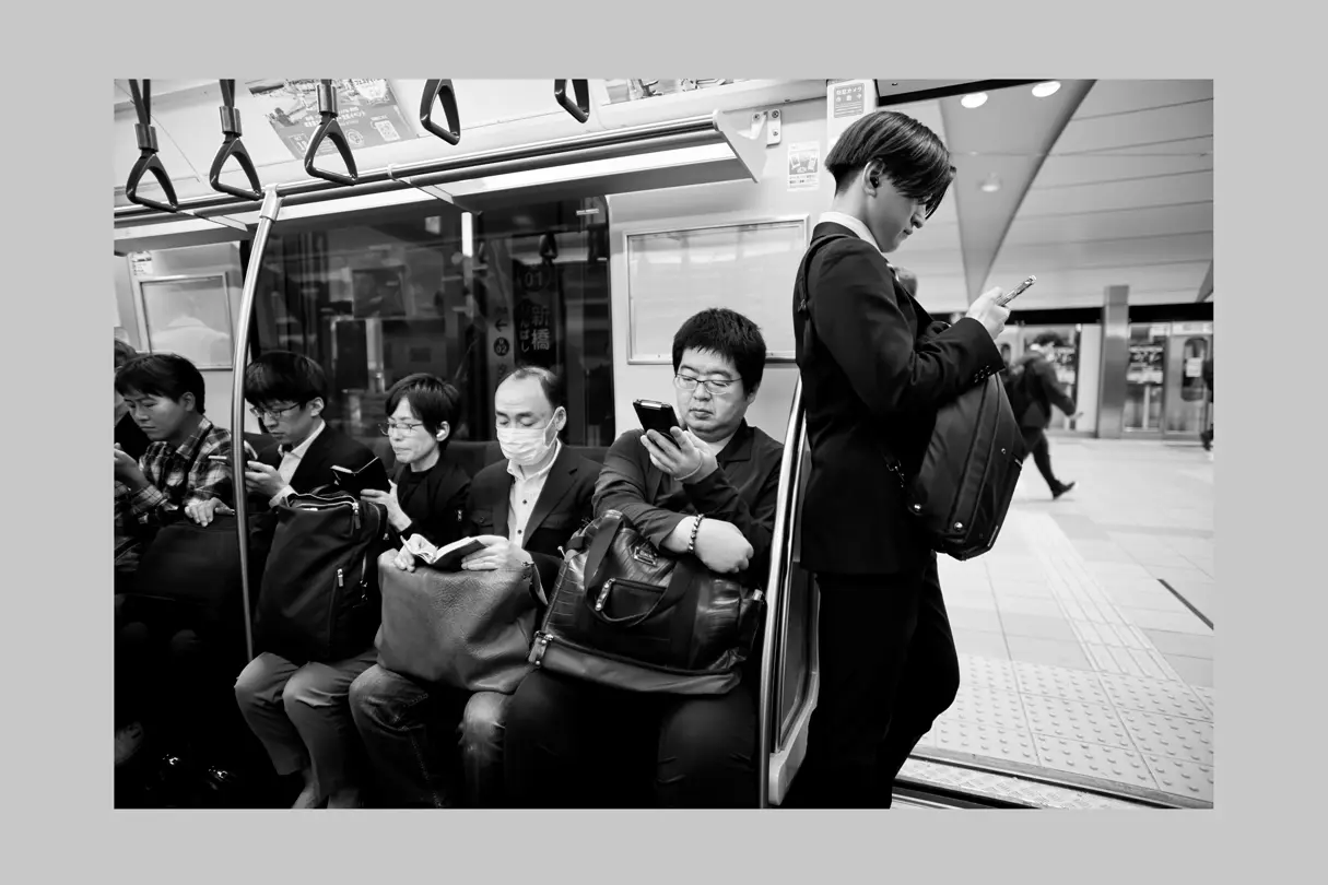 Tokyo subway commuters using smartphones during their journey, captured in black-and-white street photography.