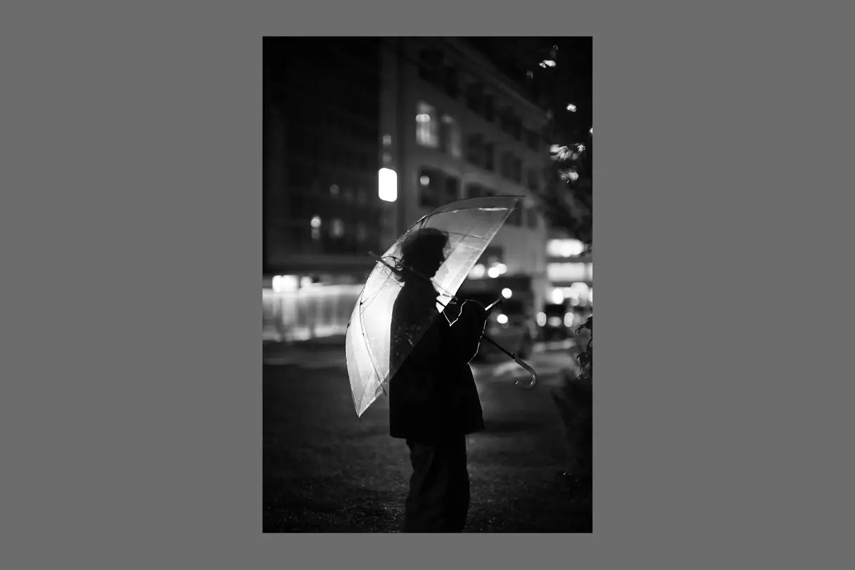 A person holding a transparent umbrella on a rainy Tokyo street at night, photographed in black and white.