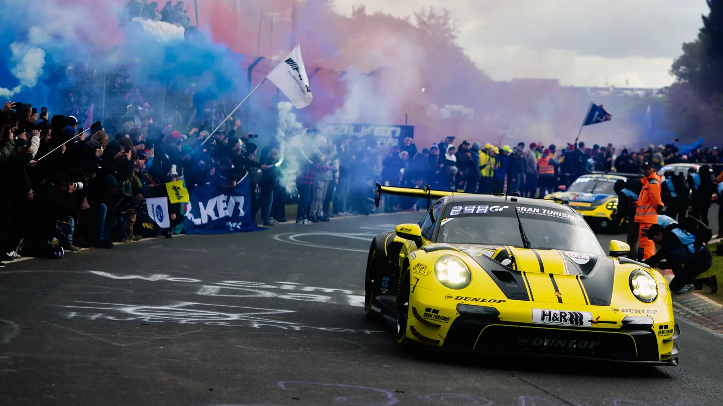 A yellow Porsche on the racetrack, with fans lining the roadside and pyrotechnics